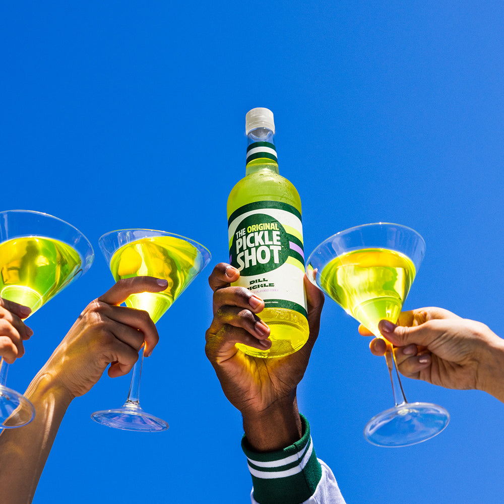 Three people clinking martini glasses of pickle cocktail around a bottle of The Original Pickle Shot against a blue sky