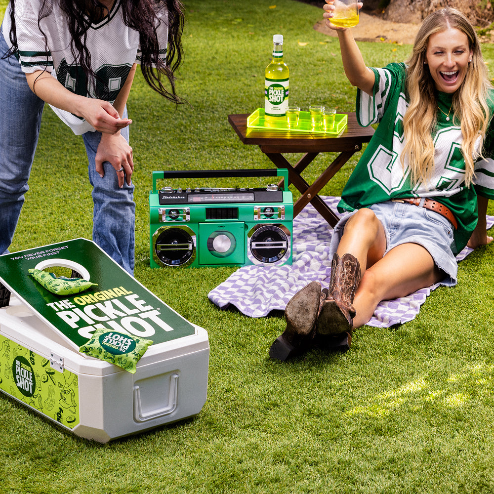 Friends sitting on the grass at a tailgate with a boombox and a cooler of The Original Pickle Shot